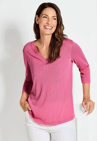 Rosa V-Ausschnitt-Oberteil mit langen Ärmeln und lockerer Passform, aus weichem Stoff gefertigt. Kombiniert mit weißen Hosen und accessorized durch die Hände in den Taschen.