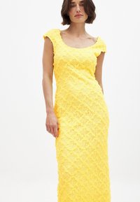 Robe en dentelle jaune avec un col rond, silhouette ajustée et motif floral texturé. Conception sans manches, style longueur sol.
