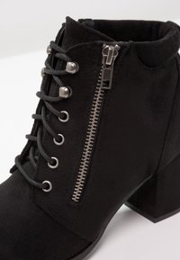 Botín de ante negro con tacón grueso, con cordones en la parte delantera, ojetes plateados y cremallera lateral para cierre. Superficie de textura lisa.