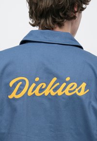 Μπλε εργασιακό μπουφάν με μυτερό γιακά, με κίτρινη κεντημένη λ έξη "Dickies" στην πλάτη. Υφασμάτινη υφή, ανθεκτικός σχεδιασμός.