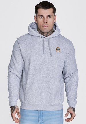 Mann med tatoveringer iført lys grå hettegenser med lite emblem på brystet og lyseblå jeans, stående mot ensfarget bakgrunn.