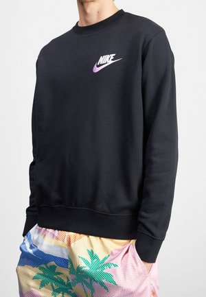 Mężczyzna w czarnej bluzie Nike z fioletowym logo, w parze z kolorowymi szortami z motywem palm.