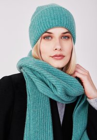 Beanie a coste color verde petrolio e sciarpa abbinata realizzate in morbido misto lana. La sciarpa si adagiate intorno al collo, mettendo in evidenza un motivo testurizzato.