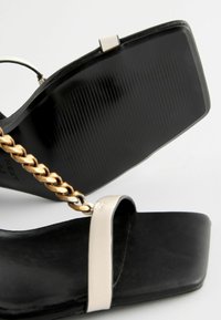Schwarze flache Sandalen mit glatter Textur, ausgestattet mit einem goldenen Kettenakzent und einem hellen Riemen; strukturierte schwarze Gummisohle.
