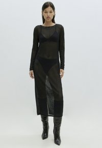 Schwarzes gestricktes Netzkleid mit langen Ärmeln, Rundhalsausschnitt und seitlichen Schlitzen; kombiniert mit schwarzen kniehohen Stiefeln mit Absätzen.