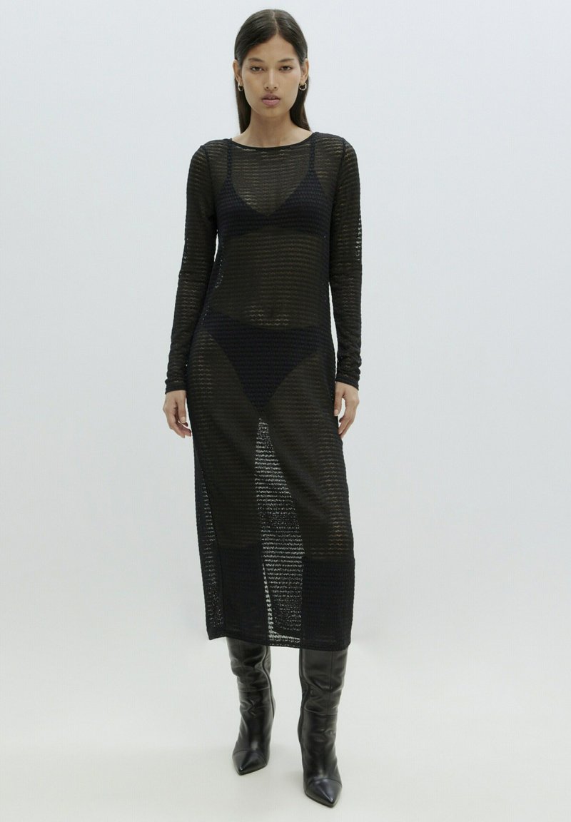 Schwarzes gestricktes Netzkleid mit langen Ärmeln, Rundhalsausschnitt und seitlichen Schlitzen; kombiniert mit schwarzen kniehohen Stiefeln mit Absätzen.