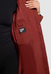 Brun quiltet jakke med en sort label på en indvendig lomme. Materialet er glat med en mat finish. Har et simpelt, rent design.
