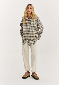 Kobieta o długich blond włosach, ubrana w dużą zielono-brązową koszulę w kratę, kremowe spodnie, czerwone skarpetki i brązowe buty turystyczne, stojąca z rękami w kieszeniach.