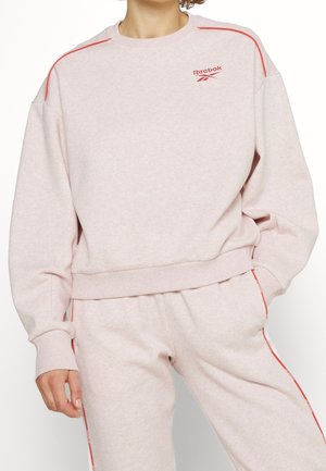 Person iført lyserød Reebok-sweatshirt og matchende bukser med røde kanter, stående med den ene hånd i lommen.