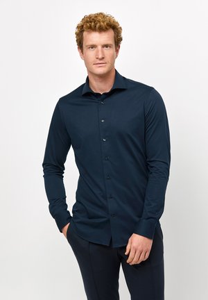 Chemise à manches longues bleu marine avec un design à boutons, dotée d'un col pointu et de boutons noirs, fabriquée à partir d'un tissu léger et lisse.