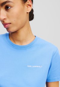 Világoskék pamut póló, kerek nyakkal, rövid ujjakkal, és fehér hímezett "KARL LAGERFELD" logóval a bal mellen.