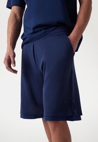 Marineblå atletiske shorts lavet af åndbart mesh-stof, med sidelommer og et diskret logo-tag i kanten. Glat finish på taljebåndet.