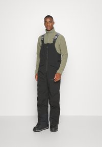 Black insulated overalls with adjustable straps and front zipper, paired with a light olive fleece top, and black sturdy winter boots.