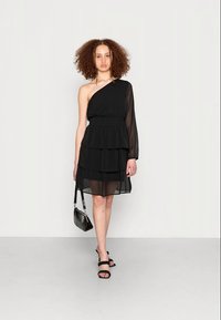 Vestido preto em camadas com um ombro, com uma cintura ajustada e mangas transparentes. Combinado com sandálias pretas e uma bolsa pequena.