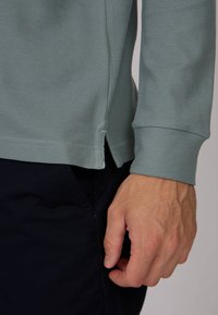Polo-shirt à manches longues vert clair en tissu texturé, avec un ourlet fendu et des poignets côtelés, porté sur un pantalon sombre.