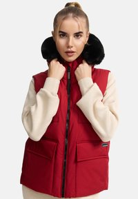Gilet rosso imbottito con collo in finta pelliccia nero, chiusura con zip e due tasche frontali. Il modello indossa un maglione beige sotto.