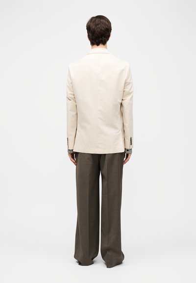 Άνδρας με σκούρα μαλλιά, γυρισμένος από την πλάτη, που φοράει κρεμ σακάκι, μανσέτες πουκαμίσου με καρό μοτίβο και φαρδιά καφέ παντελόνια, πάνω σε απλό ανοιχτόχρωμο φόντο.