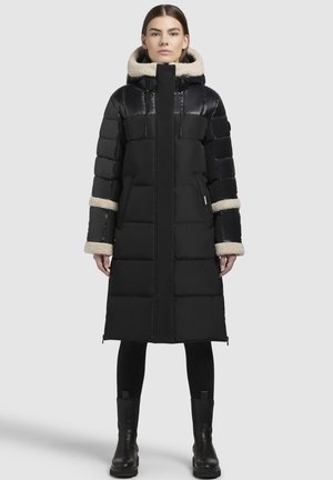 Zwarte pufferjas met een lange silhouet, glanzend bovenstuk en pluche beige capuchon. Voorzien van ribbels aan de manchetten, zijzakken en ritsen.