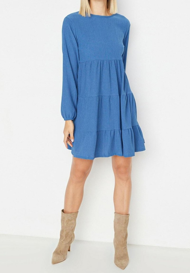 Femme portant une mini-robe bleue à manches longues et à volants, ainsi que des bottines beiges en daim à talons mi-mollet, debout devant un fond blanc.