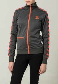 Grå zip-jacka med hög krage, med orange dragkedja och accentlinjer längs ärmarna. Mjuk, slät textur. Hummel-logg på bröstet.