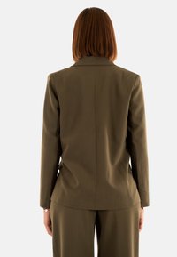 Chaqueta de traje verde olivo con cuello de muesca, mangas largas y un corte ajustado, mostrada desde la parte de atrás, exhibiendo líneas limpias y un tejido texturizado.