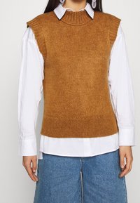 Pull sans manches marron tricoté avec un col montant et un ourlet côtelé, porté sur une chemise blanche boutonnée. Texture lisse avec une coupe décontractée.