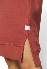 Gros plan sur l’ourlet d’un vêtement couleur rouille avec une fente latérale et une petite étiquette de marque blanche près de la fente, porté sur peau nue.