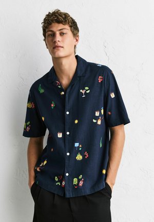 Young man with curly hair wearing a navy blue short-sleeve button-up shirt with colorful food and drink print, standing against a white wall.
