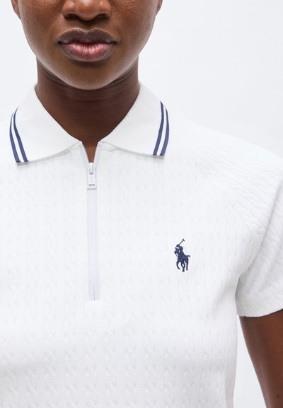 Άτομο με σκουρόχρωμο δέρμα που φοράει λευκό κοντομάνικο πόλο μπλουζάκι με διπλή μπλε ναυτικού χρώματος ρίγα στο γιακά και λογότυπο ναυτικού χρώματος στο στήθος.