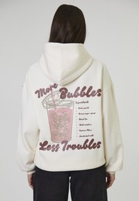 Persoon in een witte hoodie met roze bubble tea en de tekst "Meer Bubbels Minder Problemen" en een ingrediëntenlijst op de achterkant.