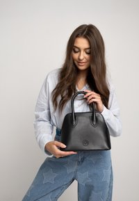 Borsa a mano in pelle nera con forma curva, doppi manici e logo in rilievo. Il modello indossa una camicia azzurra chiara e jeans con motivo a stelle.