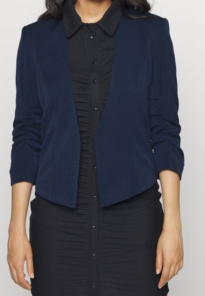 Blazer sartorial blu navy con un tessuto morbido, apertura frontale e maniche a tre quarti, indossato sopra un vestito button-up nero con colletto.