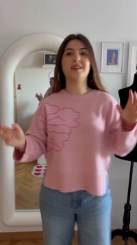 Jauna sieviete ar gariem tumšiem matiem, ģērbusies rozā džemperī un zilās džinsās, stāv pirms arkas formas spoguļa un melnas kleitas veidnes telpā.