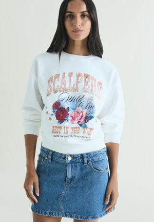 Donna con lunghi capelli scuri che indossa una felpa bianca con motivo floreale e una gonna di jeans blu, in piedi davanti a uno sfondo semplice.