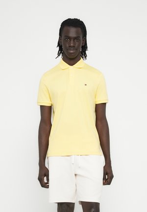 Man met dreadlocks die een geel poloshirt met korte mouwen en witte shorts met trekkoord draagt, staand tegen een effen witte achtergrond.
