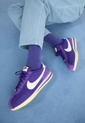 Fioletowe sneakersy Nike z białym logo, o teksturowanej powierzchni, wiązane na sznurowadła, z kontrastową kremową podeszwą, ustawione na niebieskiej powierzchni.