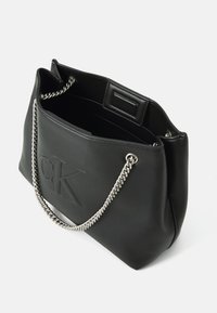 Musta nahkainen olkalaukku, jossa on kohokuvioitu "CK"-logo, hopeaketjuhihna, pehmeä tekstuuri ja avoin yläosa.