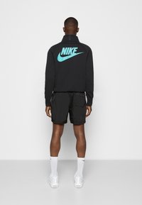 Chaqueta negra de Nike con un logotipo turquesa en la parte trasera, combinada con pantalones cortos negros y zapatillas blancas, vista desde atrás.