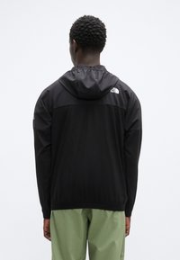 Μαύρο μπουφάν με κουκούλα και λογότυπο The North Face, με ελαφρύ σχεδιασμό και ελαστικά μανίκια. Φορεμένο πάνω από πράσινο παντελόνι, εμφάνιση από πίσω.