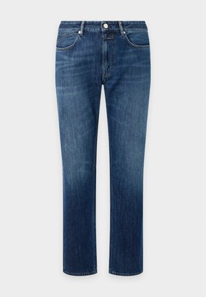 Homme debout, une main dans la poche, portant une chemise bleu marine par-dessus un T-shirt noir, un jean bleu et des baskets marron sur fond gris uni.