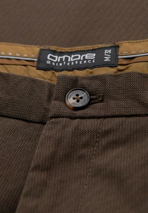 Braune Stoffhose mit strukturiertem Webmuster. Sie verfügt über einen braunen Knopf und ein Etikett mit "Ombre Quintessence" sowie der Größe M/32 auf einem sandfarbenen Bund.
