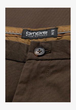 Bruna tygbyxor med en strukturerad väv. Har en brun knapp och en etikett med "Ombre Quintessence" och storlek M/32 på en ljusbrownband.