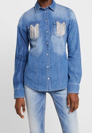 Man draagt een lichtblauw denim shirt met zilveren franjes op de borstzakken en bijpassende lichtblauwe spijkerbroek.