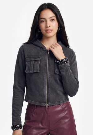 Fiatal nő hosszú sötét hajjal sötétszürke cipzáras kapucnis pulóvert visel zsebekkel és szegecselt csuklópántokkal, bordó bőrnadrággal párosítva.