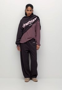 Hanorac larg negru cu text roz suprapus peste un tricou maro cu dungi și mâneci lungi, asortat cu pantaloni sport negri largi și adidași bej.