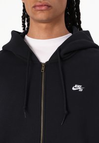 Schwarze Zip-Hoodie aus Baumwollmischung, ausgestattet mit einem goldenen Reißverschluss, einer Kordel und kleinem weißen Nike SB-Logo auf der linken Brust.