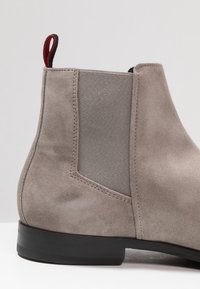 Bottines Chelsea en daim gris clair avec panneau élastique noir sur le côté, semelle noire et tirant rouge et noir à l'arrière.