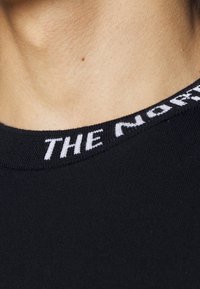Gros plan sur le cou et le haut de la poitrine d'une personne portant une chemise noire avec un logo blanc "THE NORTH" brodé près du col.