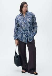 Camicia a fantasia paisley blu navy con chiusura a pulsante, abbinata a pantaloni larghi marroni e a una borsa a tracolla nera intrecciata. Texture liscia.