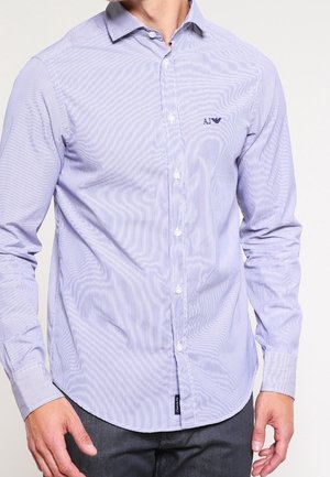 Mężczyzna ubrany w jasnoniebieską koszulę w paski z długim rękawem i czarne spodnie, logo marki na lewej piersi.